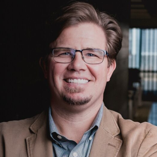 A smiling man, Bryce H. Chapman n in a tan blazer and glasses stands with arms crossed in a hallway, leaning against a dark wall with soft lighting.