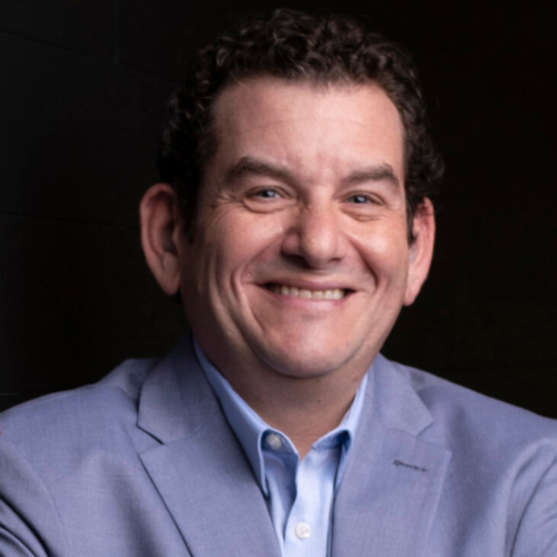 A man, Evan Winegard in a light purple blazer and light blue shirt stands smiling with arms crossed, leaning against a dark wood-paneled wall. The lighting emphasizes his confident, friendly expression, and the background gradually fades into darkness.
